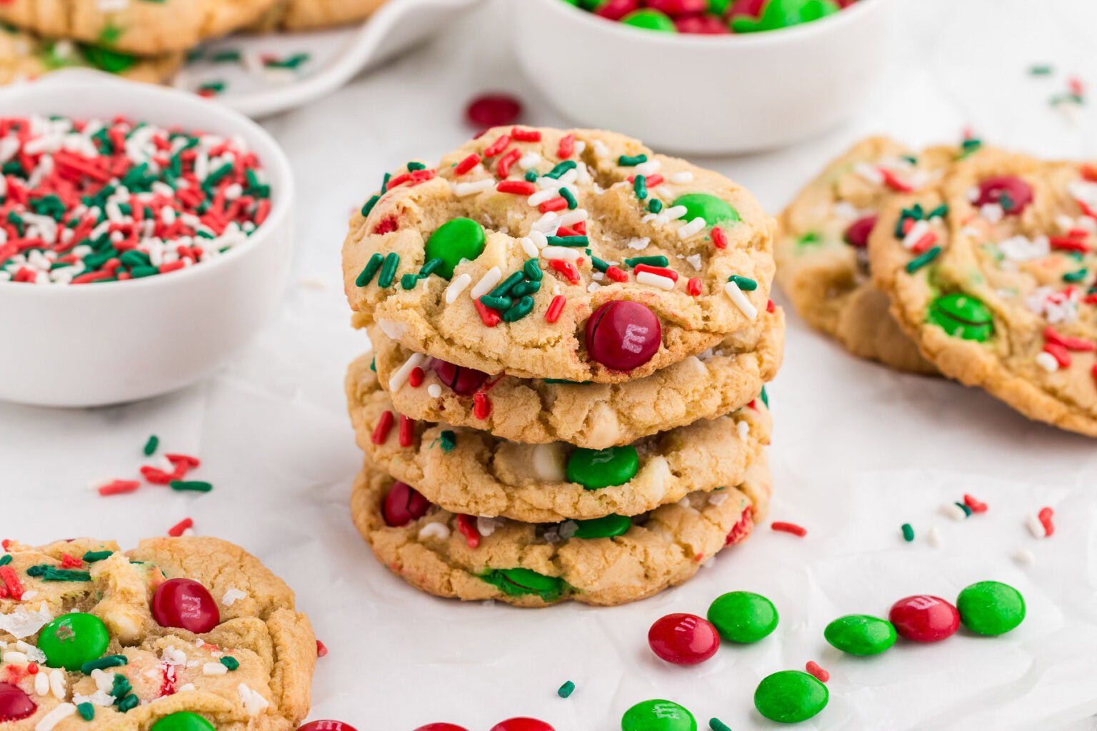 A stack of festive cookies with red, green, and white chocolate candies and sprinkles, surrounded by bowls of sprinkles and loose candies on a white surface.