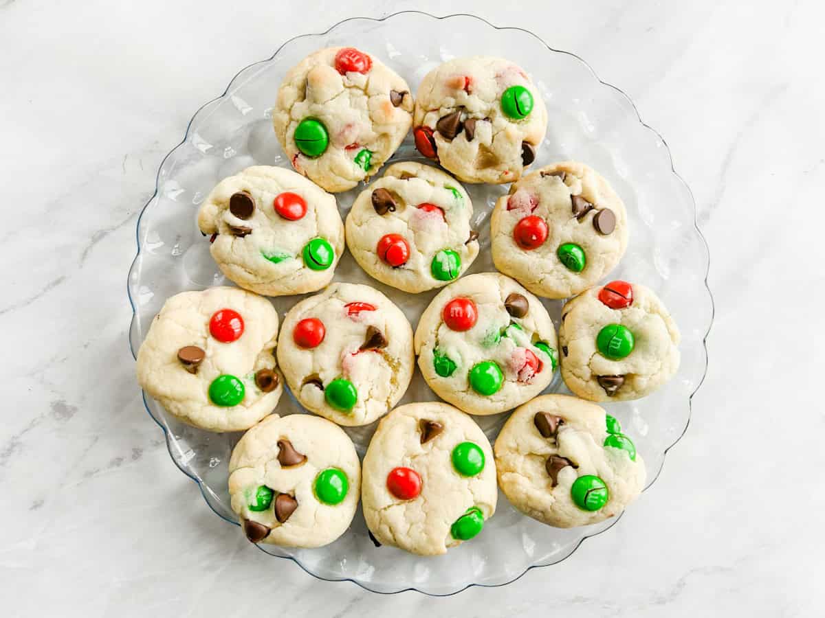 A glass plate holding a batch of cookies topped with red and green chocolate candies and chocolate chips, arranged on a white surface.