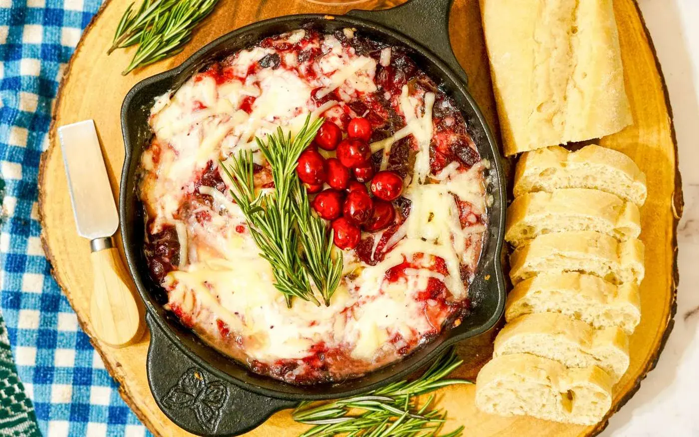 Baked brie topped with cranberries, rosemary, and melted cheese in a skillet, served with sliced baguette on a wooden board.