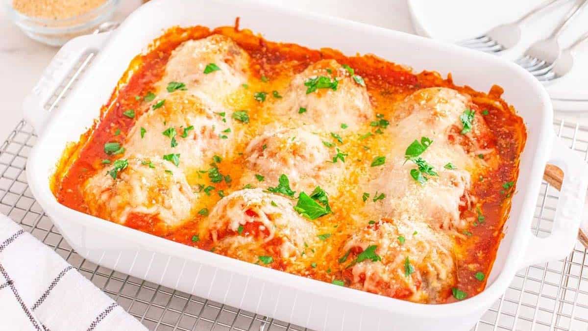 A white baking dish with baked chicken breasts topped with tomato sauce, melted cheese, and chopped parsley, placed on a cooling rack.