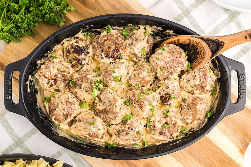A black oval dish filled with meatballs in a creamy sauce, garnished with chopped parsley, with a wooden spoon resting inside.