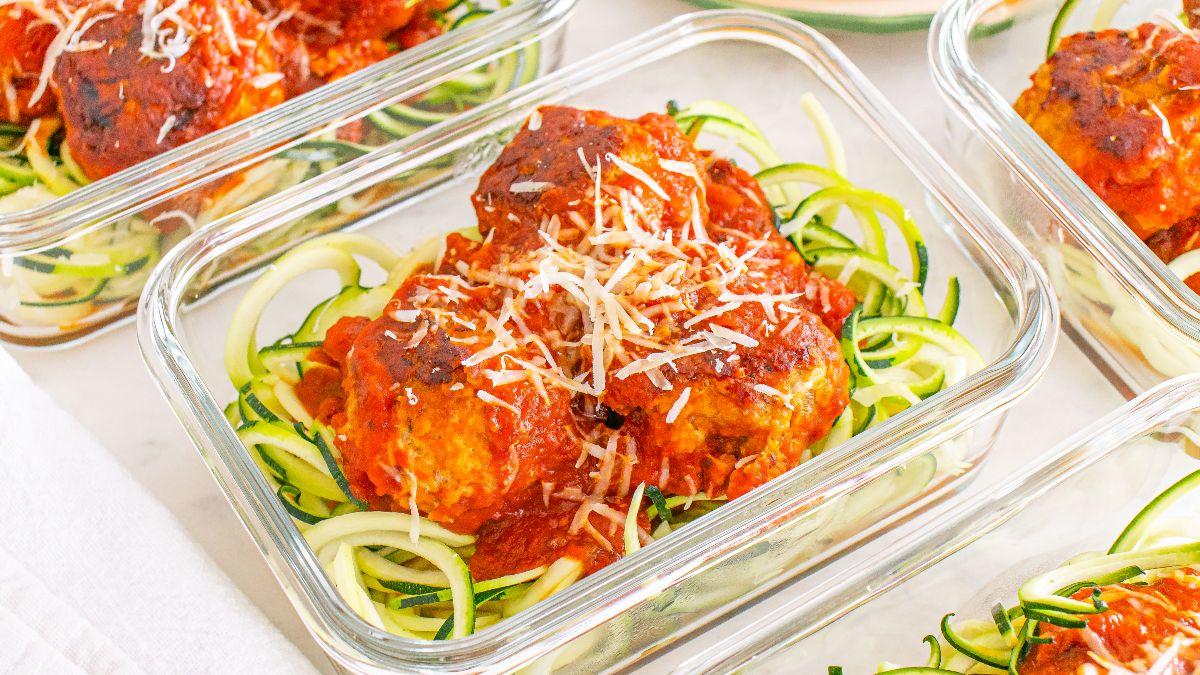 Glass containers filled with zucchini noodles topped with meatballs, marinara sauce, and shredded cheese.
