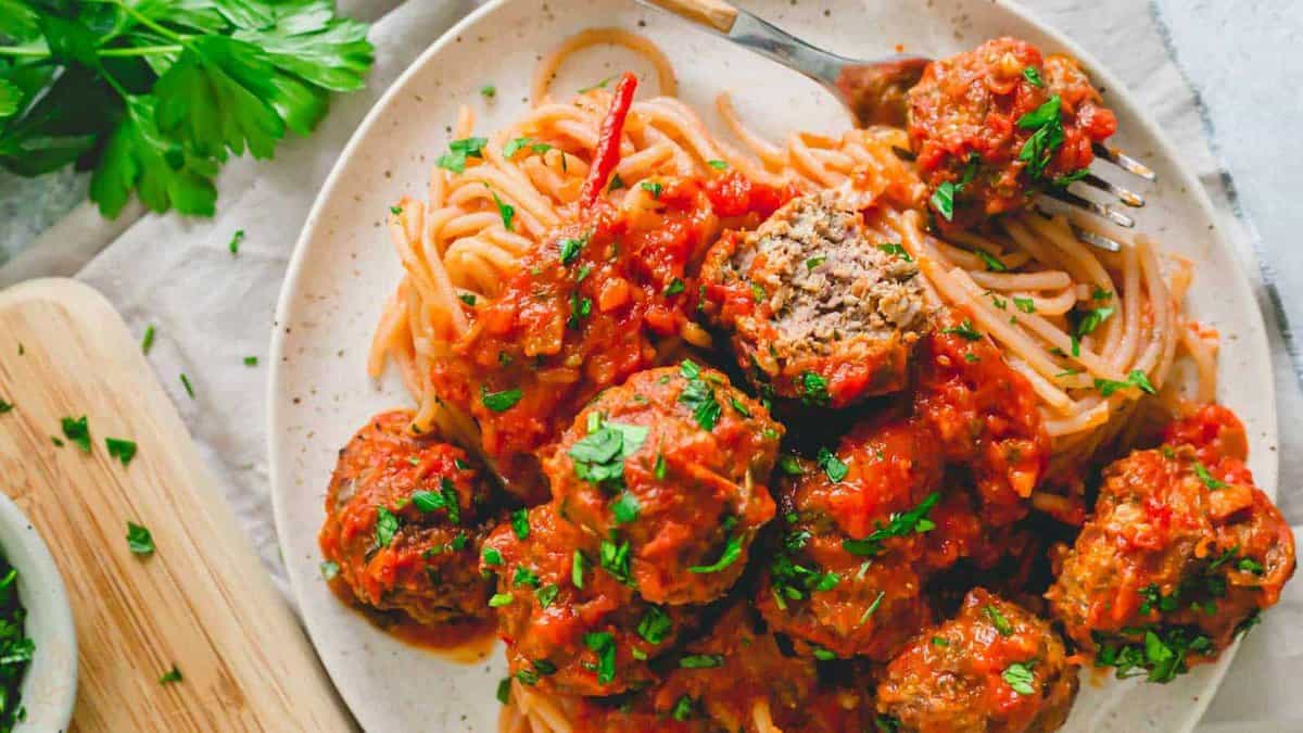 A plate of spaghetti topped with meatballs in tomato sauce, garnished with chopped parsley, with a fork and fresh herbs on the side.