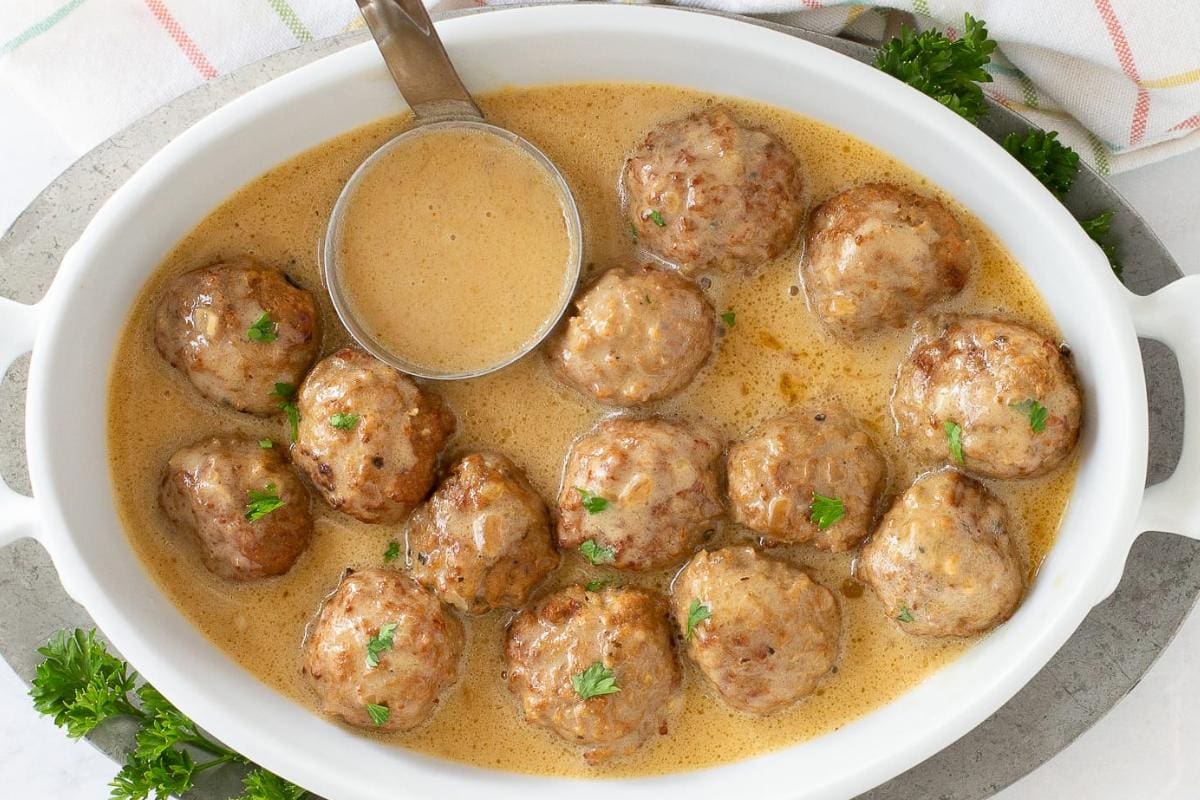 A white oval dish filled with meatballs in creamy brown sauce, garnished with parsley, with a metal ladle resting inside.