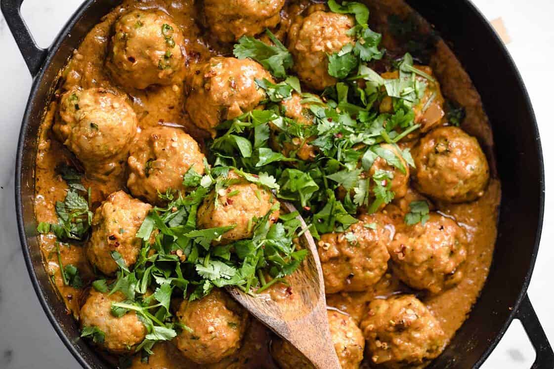 Meatballs in a creamy, spiced sauce garnished with chopped cilantro, served in a black skillet with a wooden spoon.