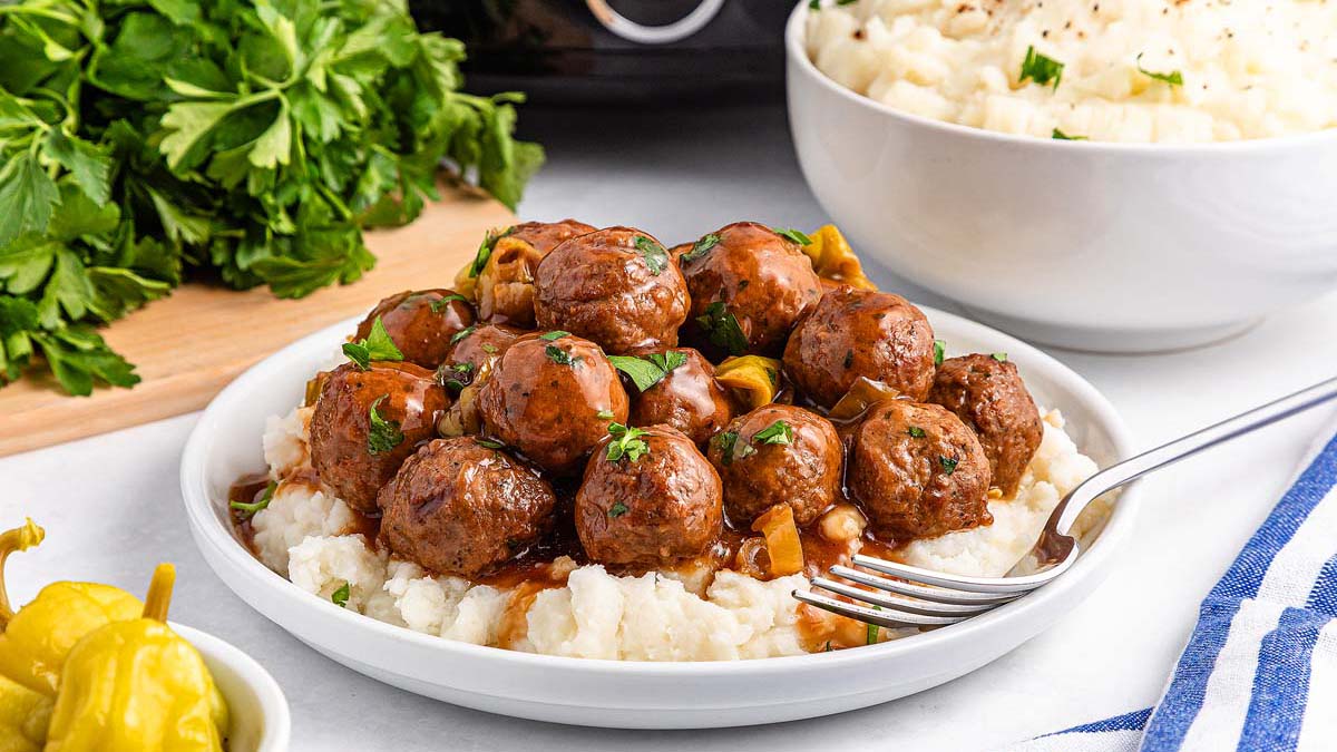 A plate of mashed potatoes topped with meatballs in brown gravy, garnished with chopped parsley, sits beside a bowl of mashed potatoes and fresh parsley.