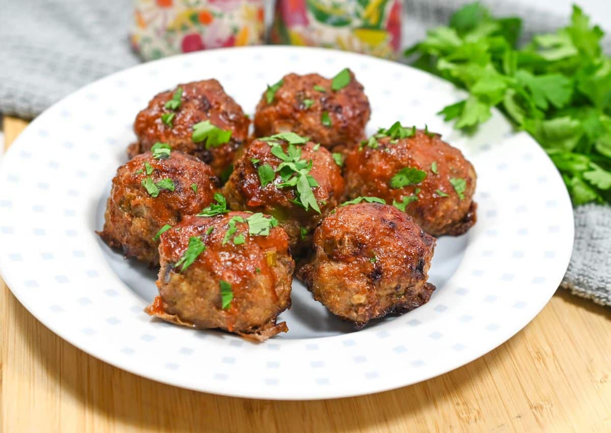 Seven meatballs garnished with chopped parsley are arranged on a white plate, placed on a wooden surface next to a bunch of fresh parsley.