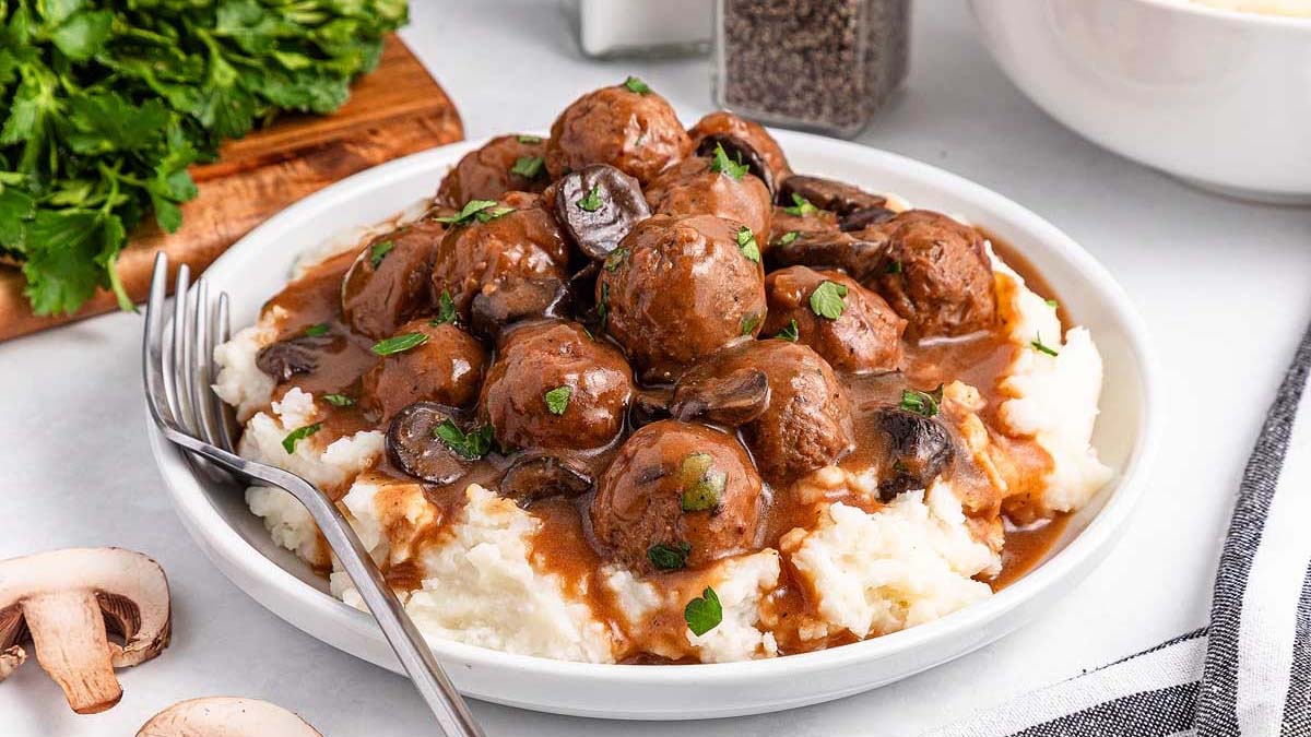 A plate of mashed potatoes topped with meatballs, brown gravy, and mushrooms, garnished with chopped parsley; a fork rests on the plate.