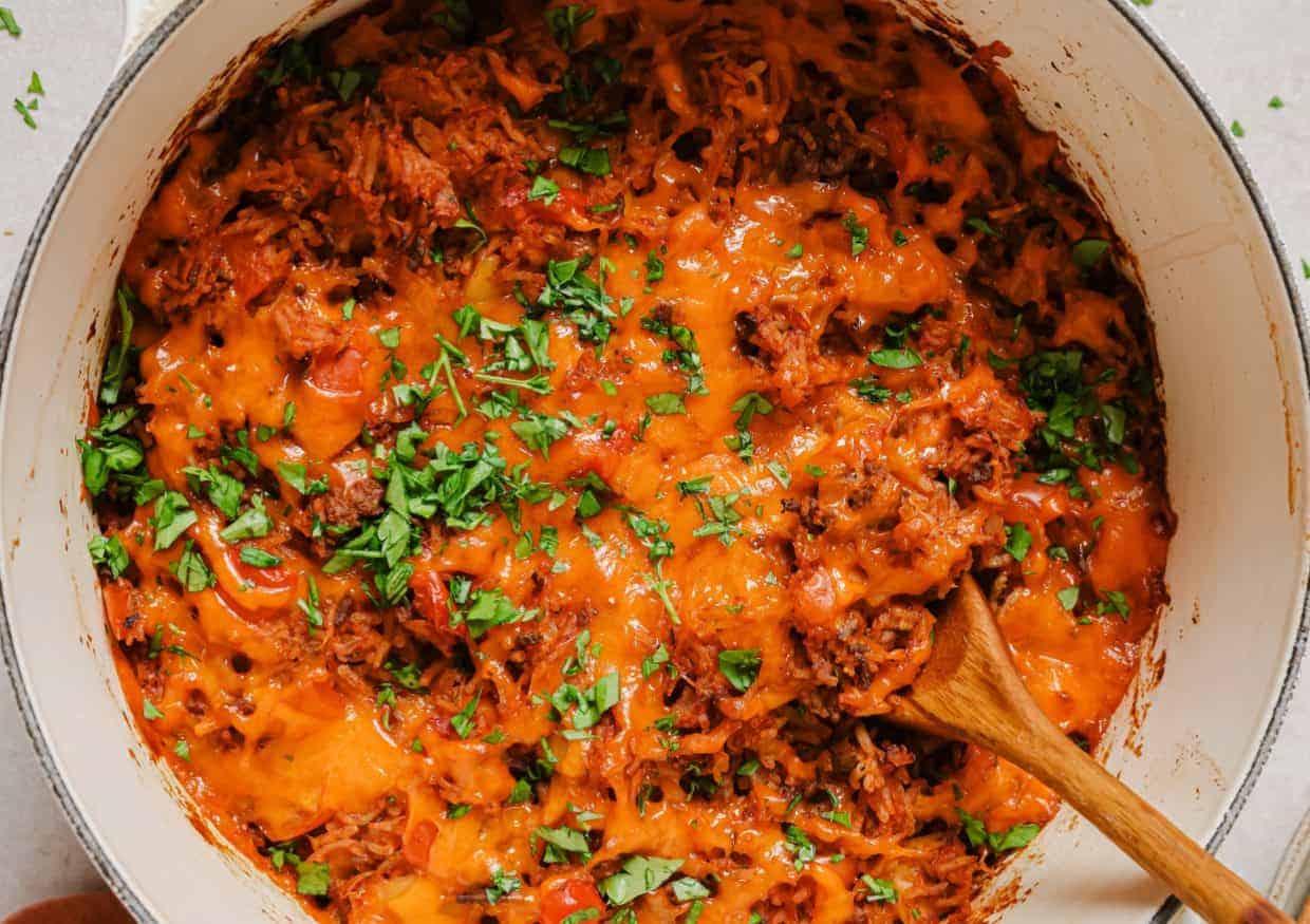 A pot filled with shredded beef, melted cheese, chopped tomatoes, and herbs, with a wooden spoon resting inside.