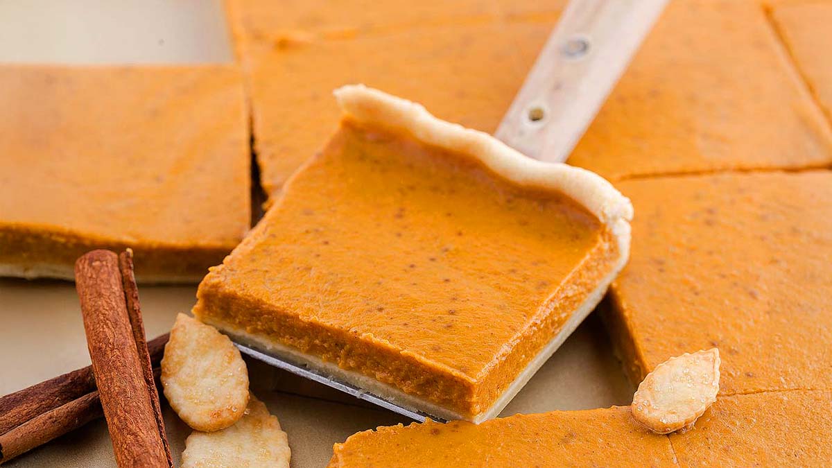 A square slice of pumpkin pie bar with a flaky crust on a spatula, surrounded by additional bars, cinnamon sticks, and pie crust decorations.