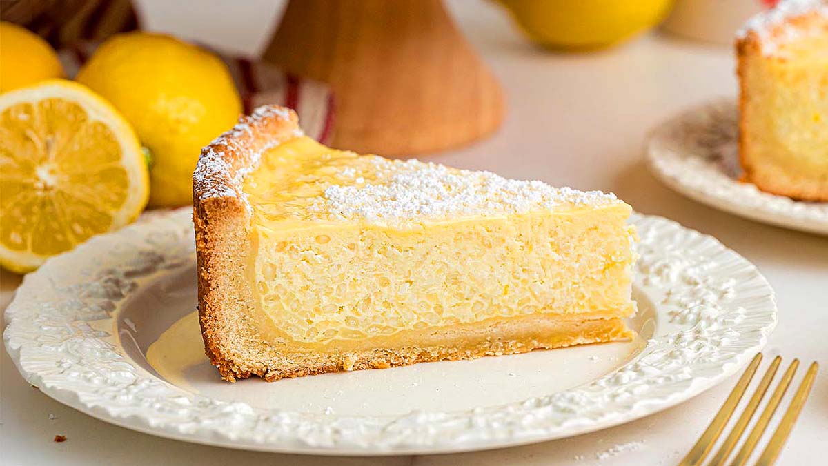 A slice of lemon ricotta pie dusted with powdered sugar sits on a white plate, with fresh lemons in the background.