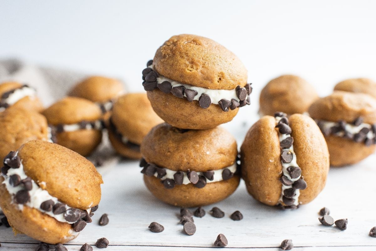 Pumpkin whoopie pies filled with cream and rolled in chocolate chips, stacked and scattered on a white wooden surface.