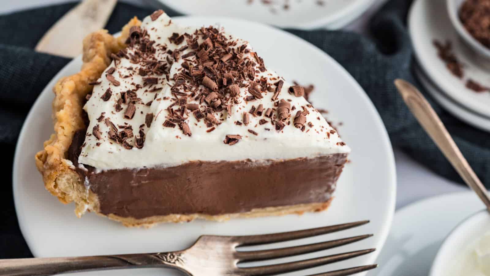 A slice of chocolate cream pie topped with whipped cream and chocolate shavings on a white plate with a fork.