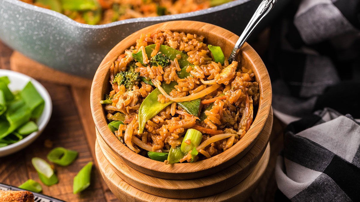 A wooden bowl filled with vegetable fried rice, including broccoli, snap peas, and carrots, with a spoon inside. A skillet and sliced green onions are visible in the background.