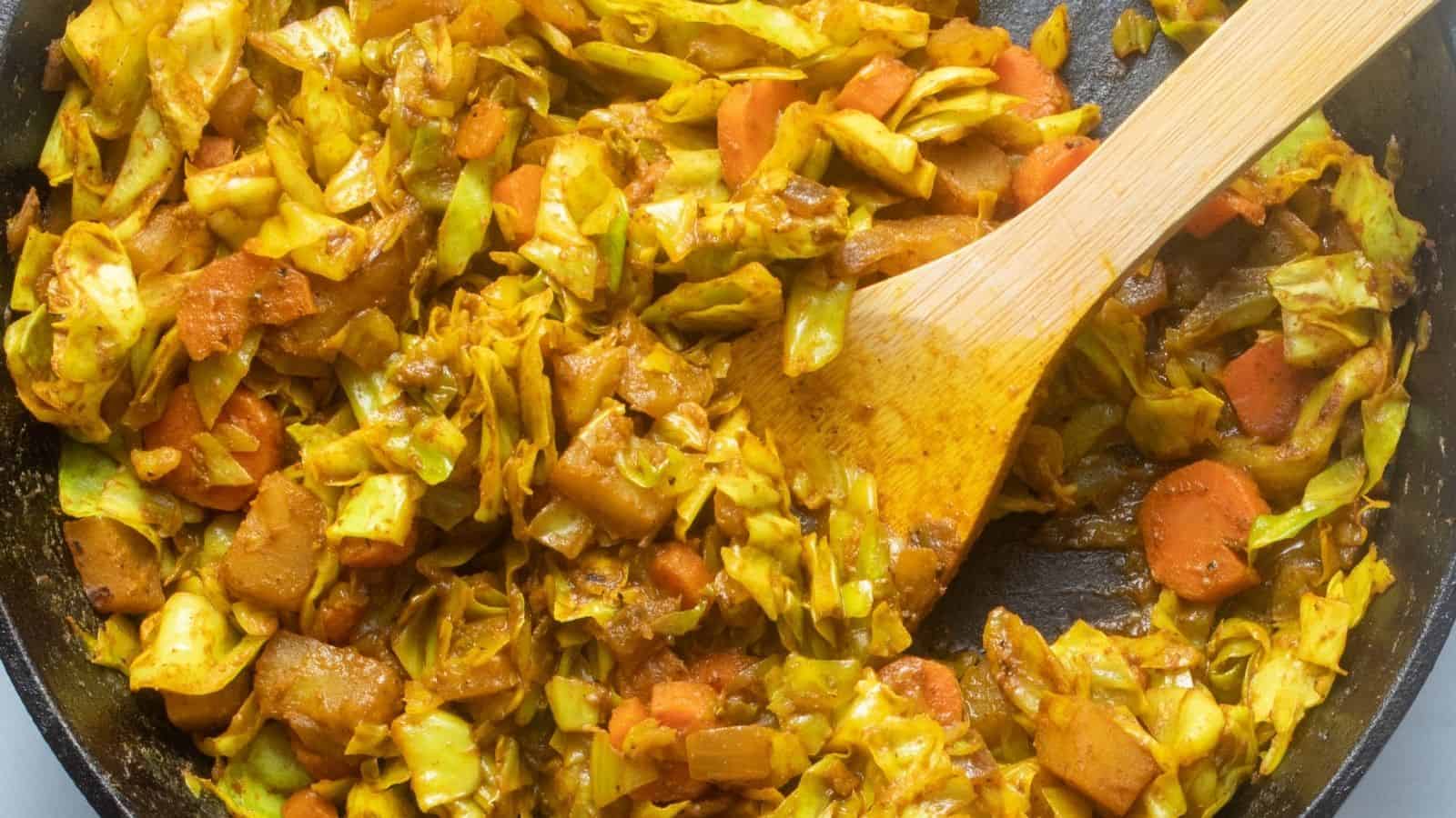 A close-up of a pan filled with sautéed cabbage, carrots, and potatoes, being stirred with a wooden spatula.