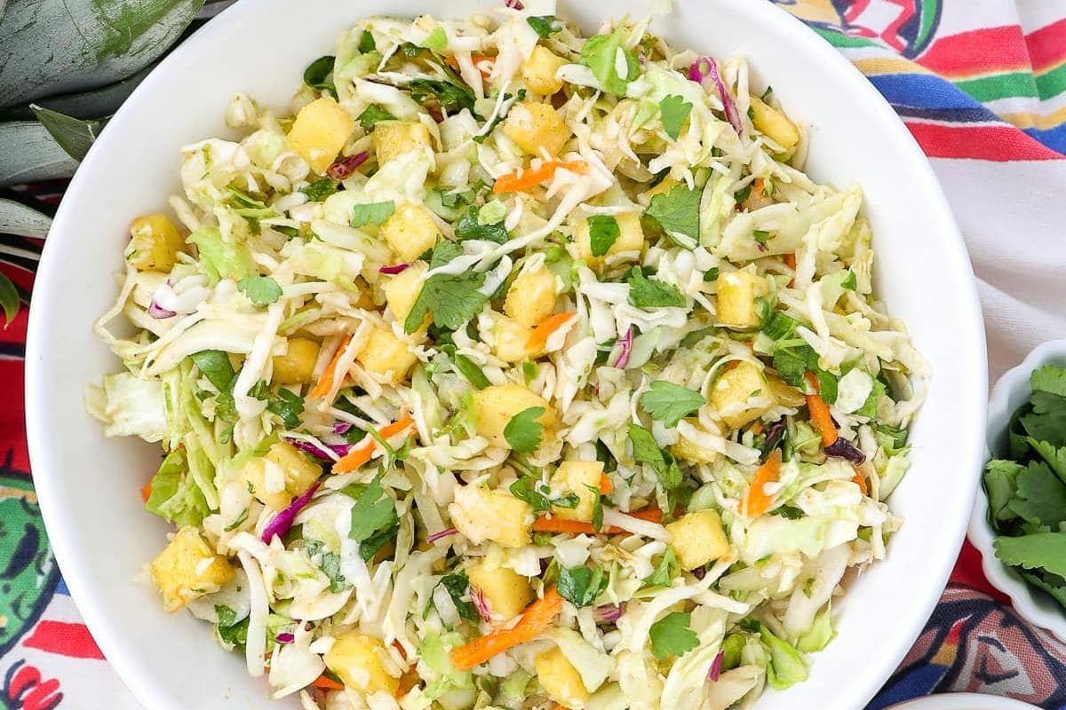 A bowl of cabbage slaw with pineapple chunks, shredded carrots, cilantro, and other vegetables on a colorful cloth background.