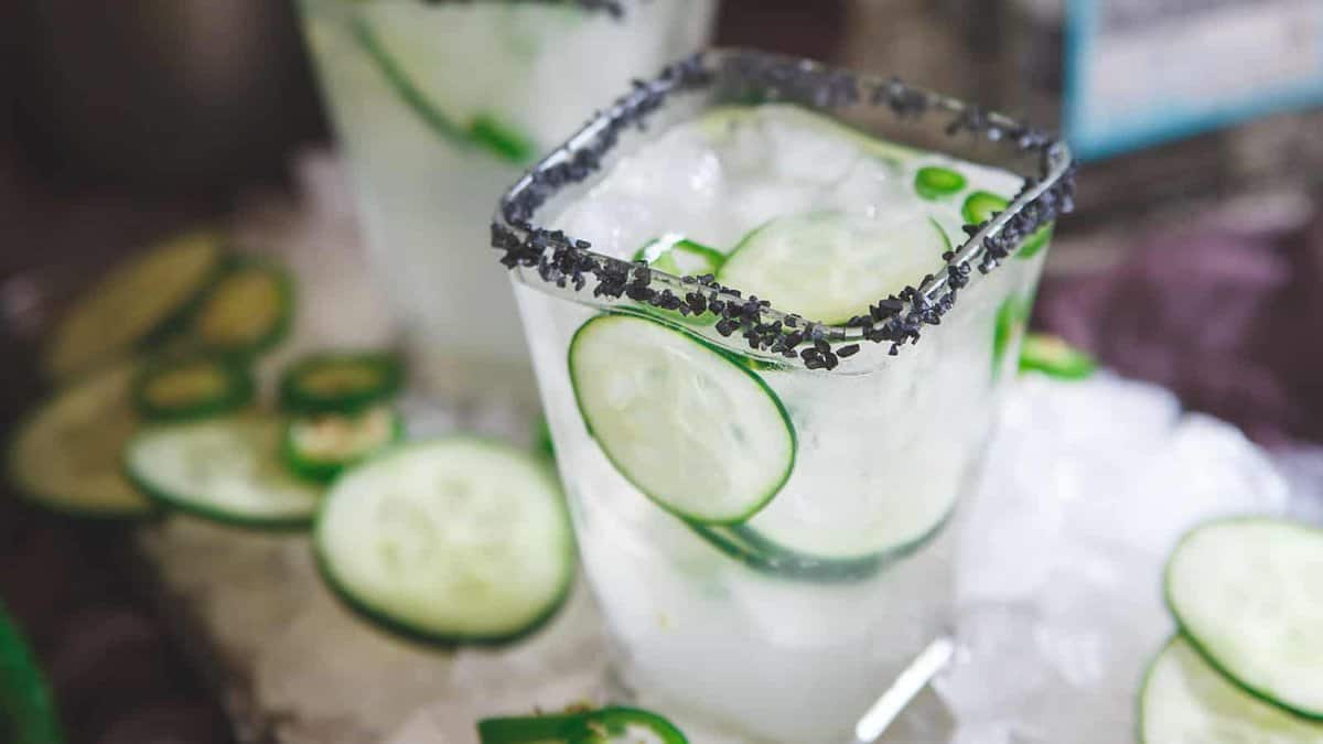 A glass of iced cocktail with cucumber slices and a black salt rim sits on a bed of ice, with extra cucumber slices nearby.