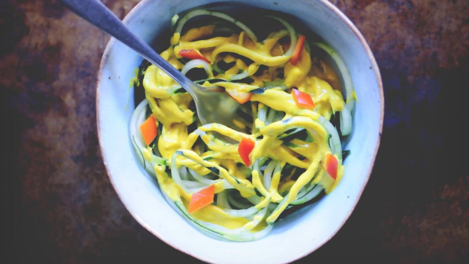 A bowl of spiralized vegetables topped with yellow sauce and diced red peppers, with a fork resting inside.