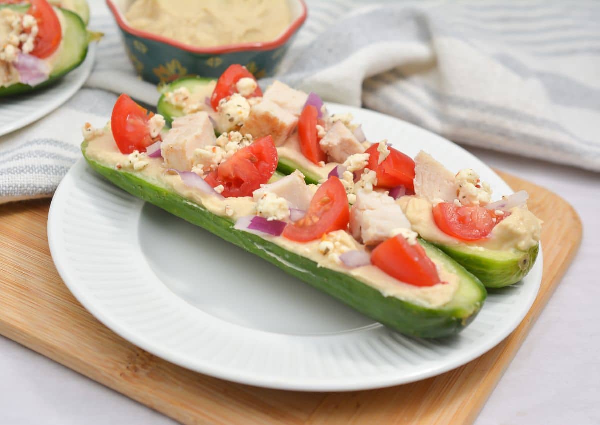 A halved cucumber filled with hummus, diced chicken, tomato slices, red onion, and crumbled cheese on a white plate.
