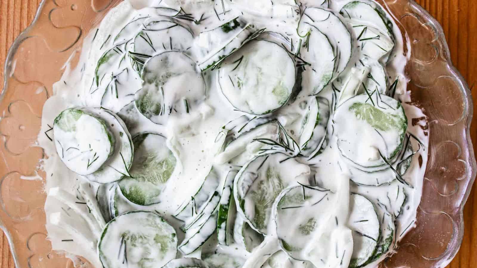 A glass bowl filled with sliced cucumbers and onions coated in a creamy white dressing, garnished with fresh dill.