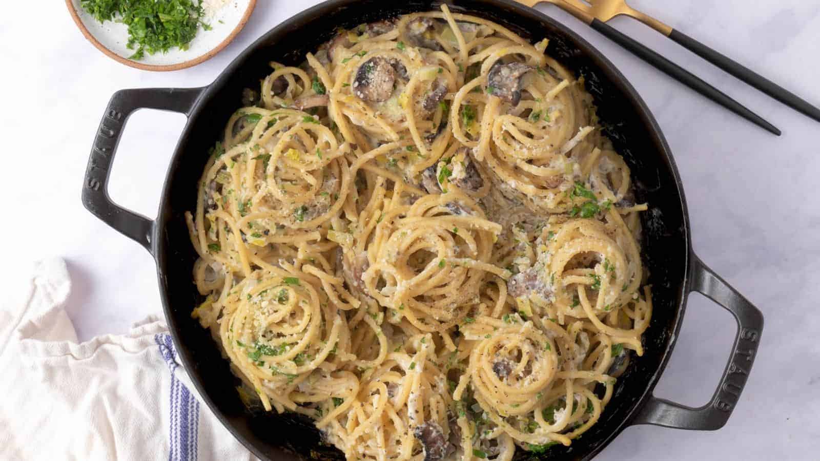 A skillet filled with cooked spaghetti mixed with a creamy sauce, mushrooms, and chopped herbs, with a plate of herbs and utensils nearby.