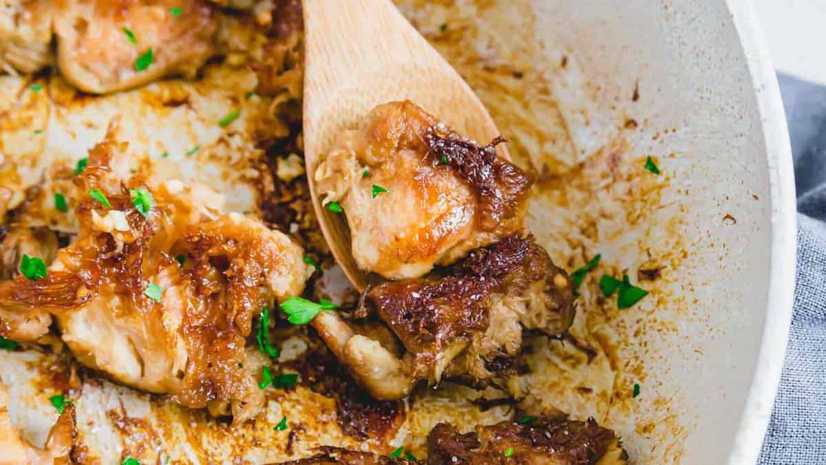 Close-up of seared chicken pieces in a skillet, garnished with chopped herbs, with a wooden spoon lifting a portion.
