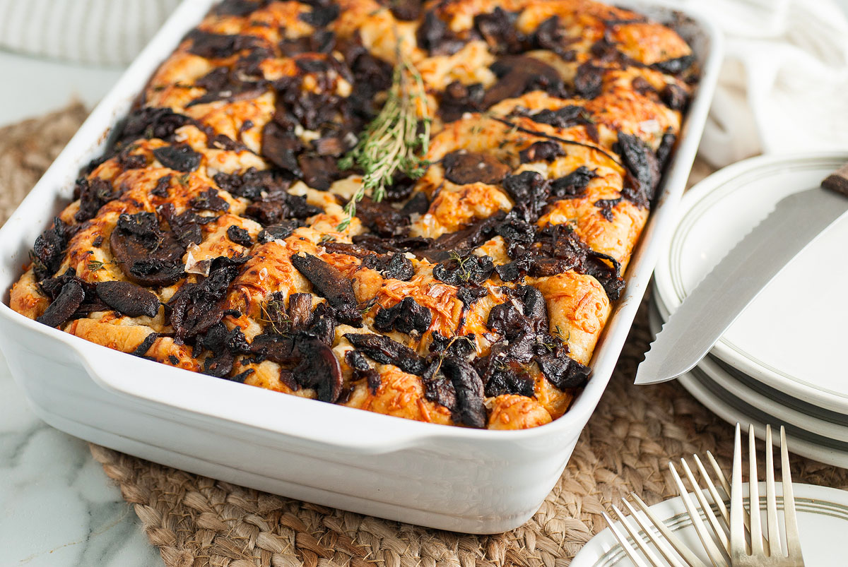 A baked casserole topped with browned mushrooms and herbs in a white dish, placed beside stacked plates, a knife, and forks on a woven mat.