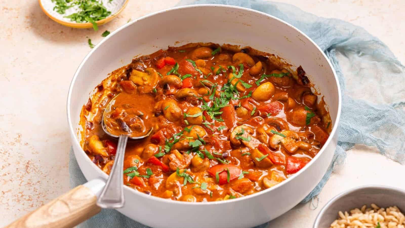 A pan filled with mushroom and bell pepper stew in a tomato-based sauce, garnished with chopped herbs, with a spoon resting inside.