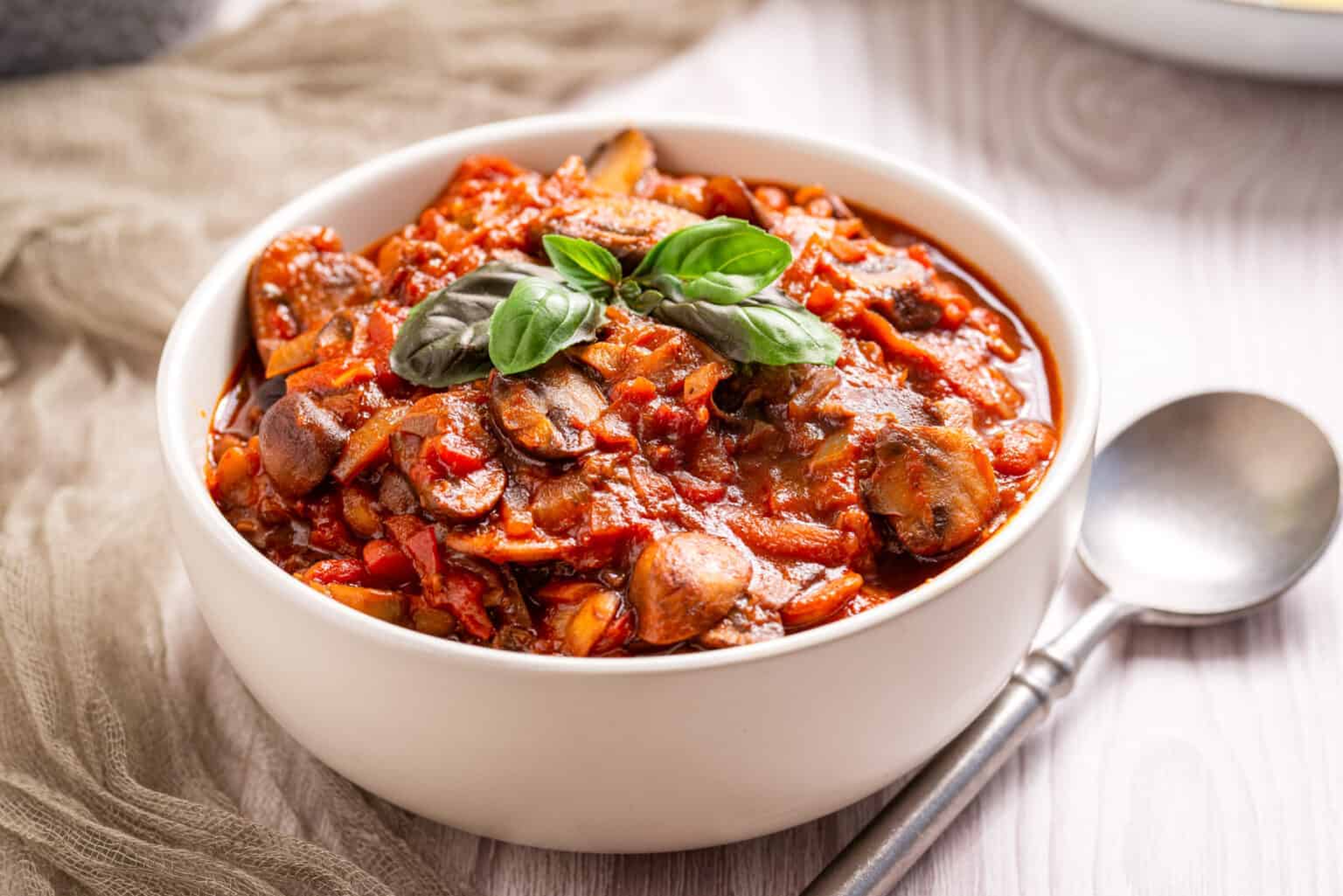 A white bowl filled with mushroom and tomato stew, garnished with fresh basil leaves, sits beside a spoon on a light wooden surface.
