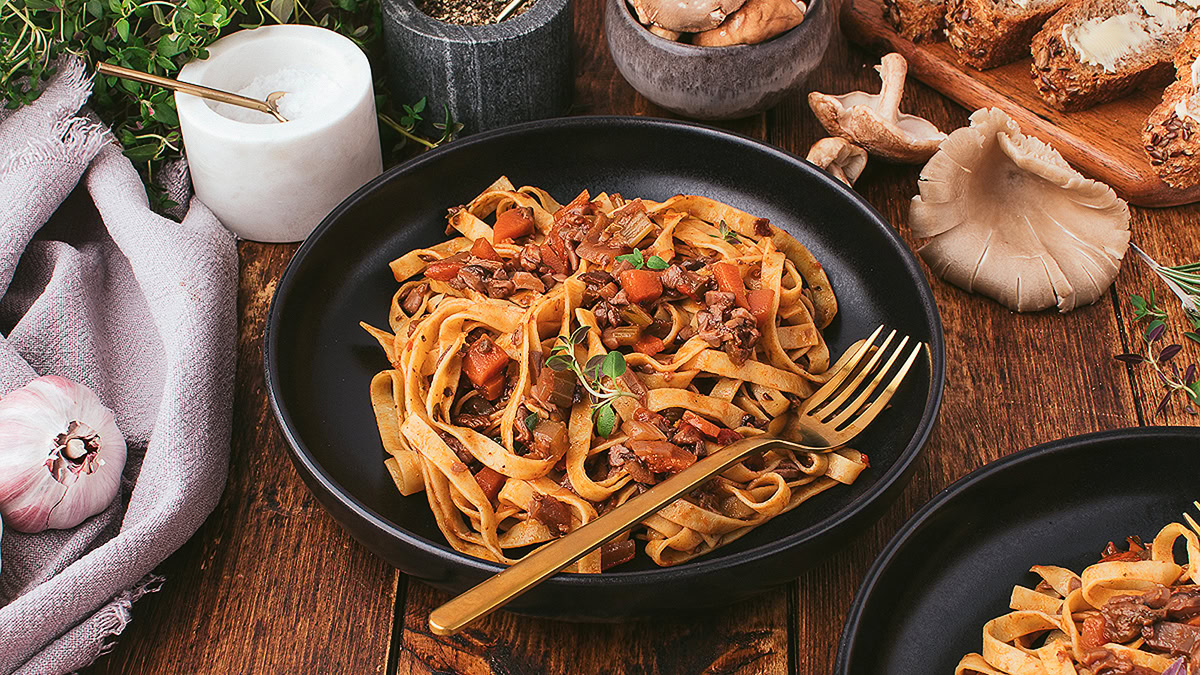 A black bowl of tagliatelle pasta with a mushroom and tomato sauce sits on a wooden table, garnished with herbs and served with a gold fork. Other ingredients surround the bowl.