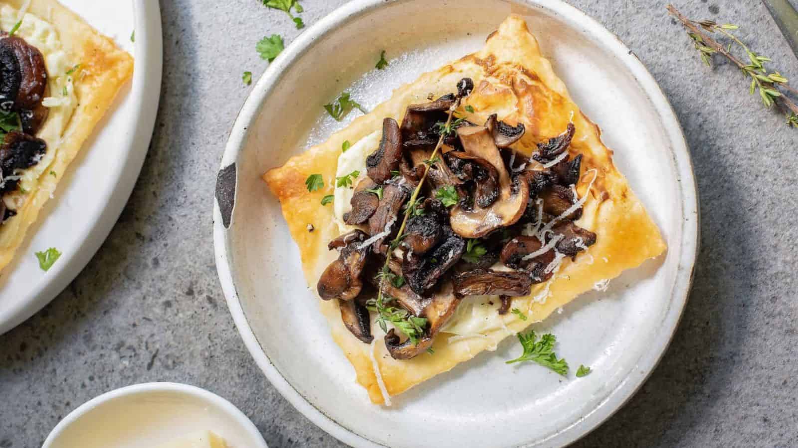 A square piece of puff pastry topped with sautรฉed mushrooms and herbs is served on a round white plate.