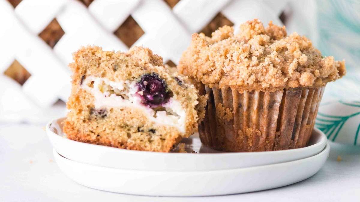 A whole crumb-topped muffin and a halved muffin with a cream cheese and blueberry filling are placed on a white plate.