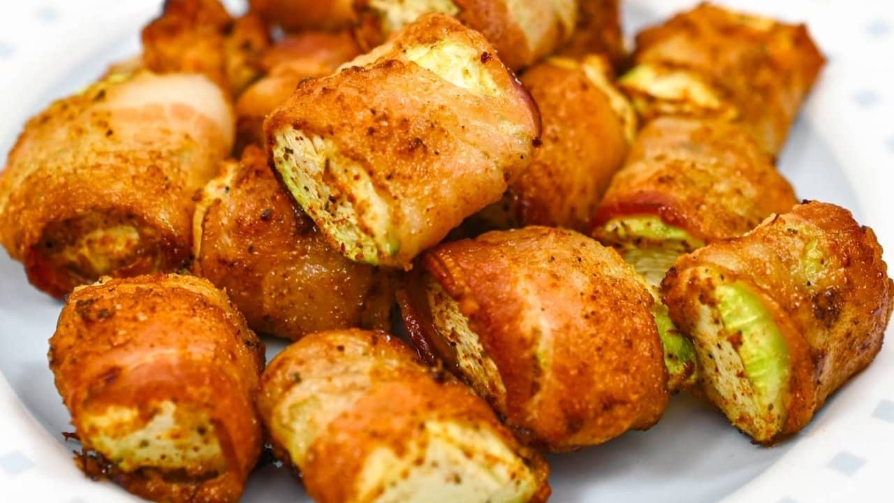 Close-up of several pieces of cooked bacon-wrapped zucchini on a white plate.