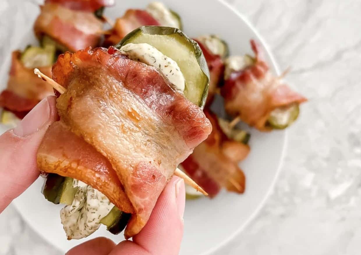 A hand holds a bacon-wrapped pickle slice filled with a creamy herb mixture, secured with a toothpick. Other similar appetizers are on a white plate in the background.
