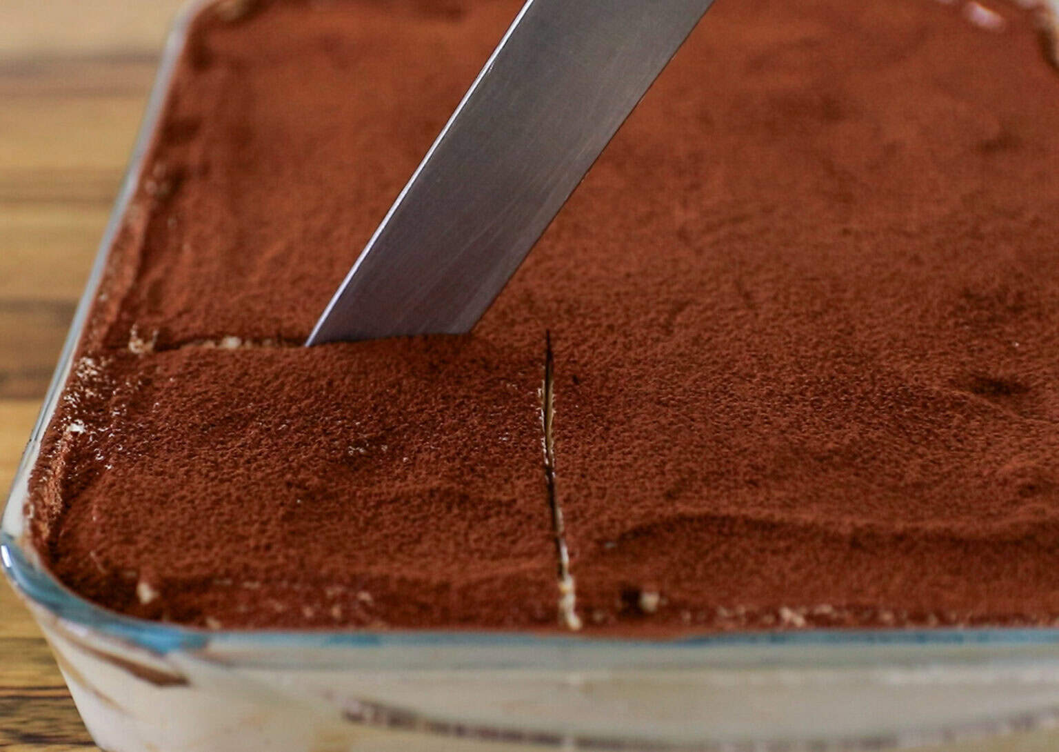 A knife is cutting into a rectangular dish of tiramisu topped with cocoa powder.