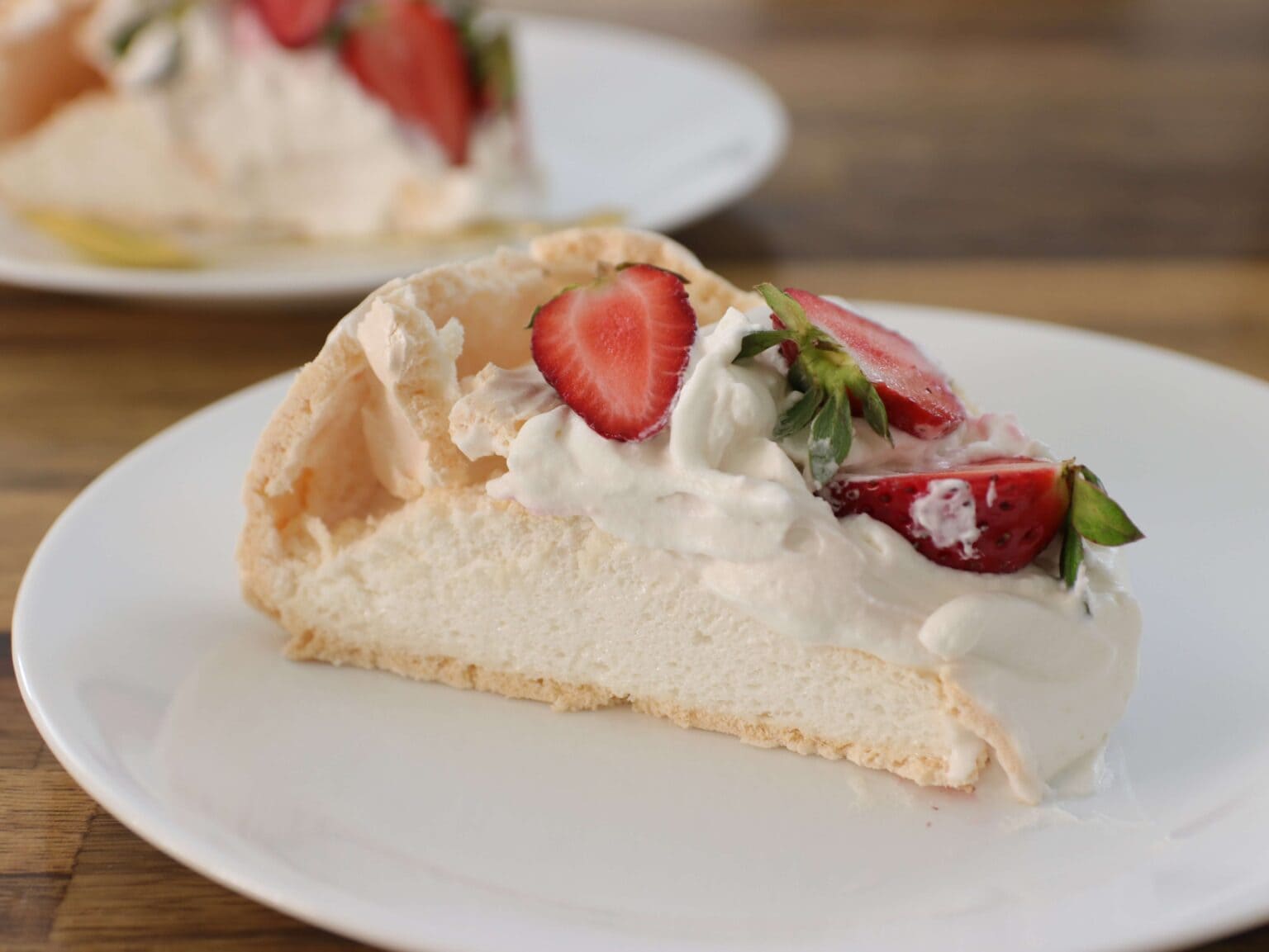 A slice of pavlova topped with whipped cream and sliced strawberries on a white plate, with another slice blurred in the background.