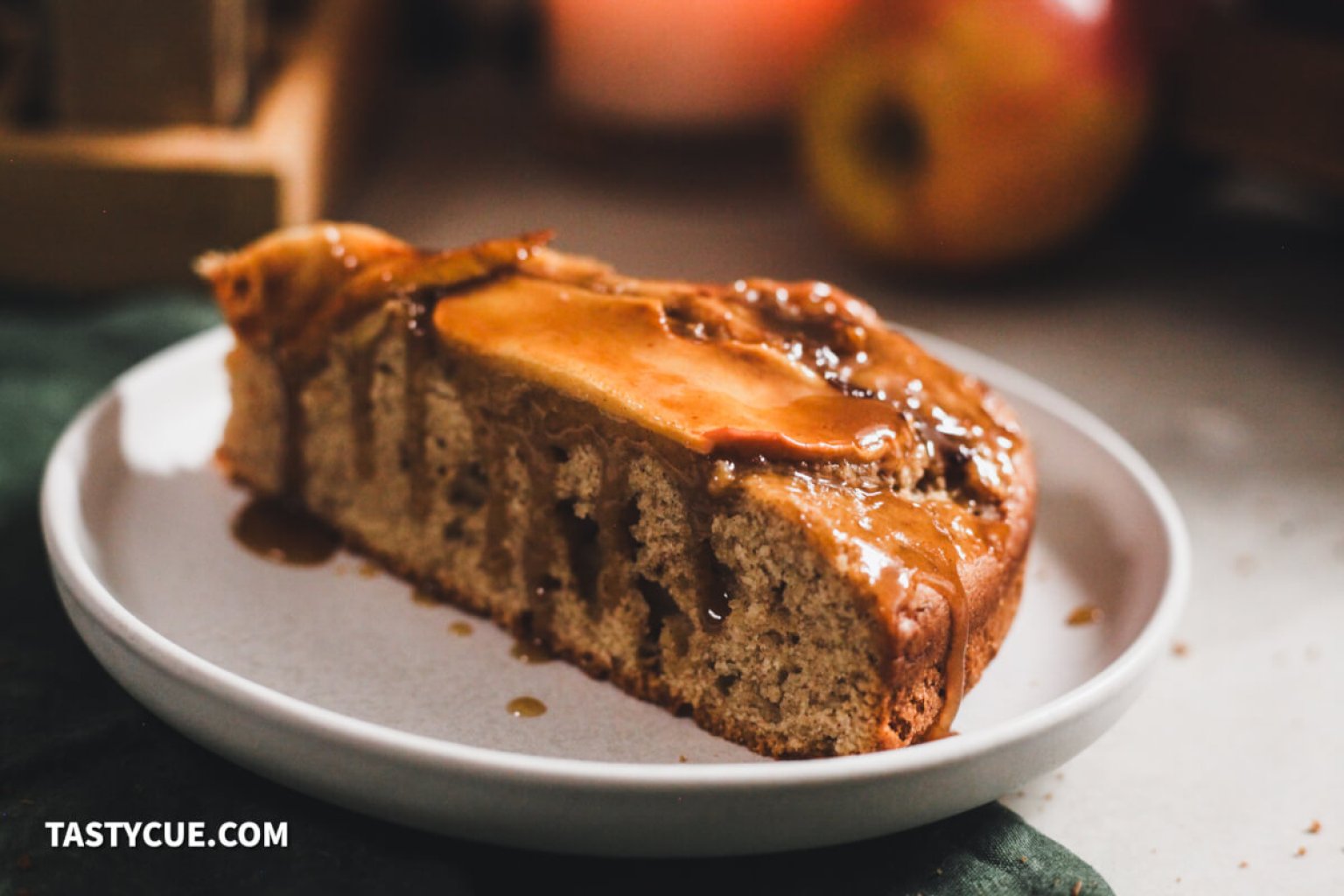 A slice of apple cake topped with caramel sauce sits on a white plate, with an apple and blurred background objects nearby.