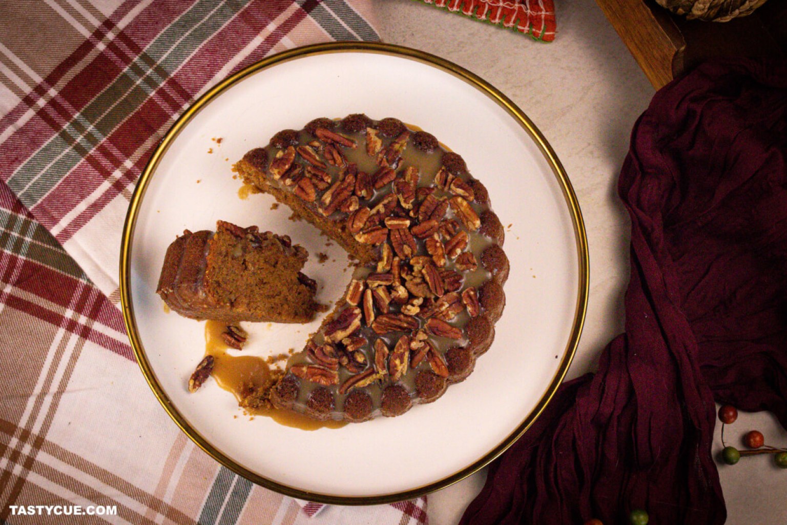 A round pecan-topped cake with caramel sauce sits on a white plate; one slice is cut and slightly removed from the cake. A plaid cloth and red napkin are nearby.
