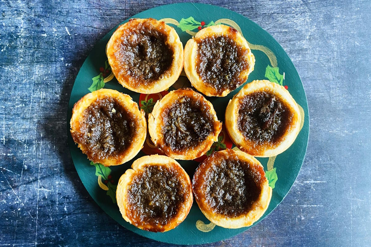 Eight butter tarts with golden crusts and dark filling arranged in a circle on a green, floral-patterned plate, set on a textured blue surface.