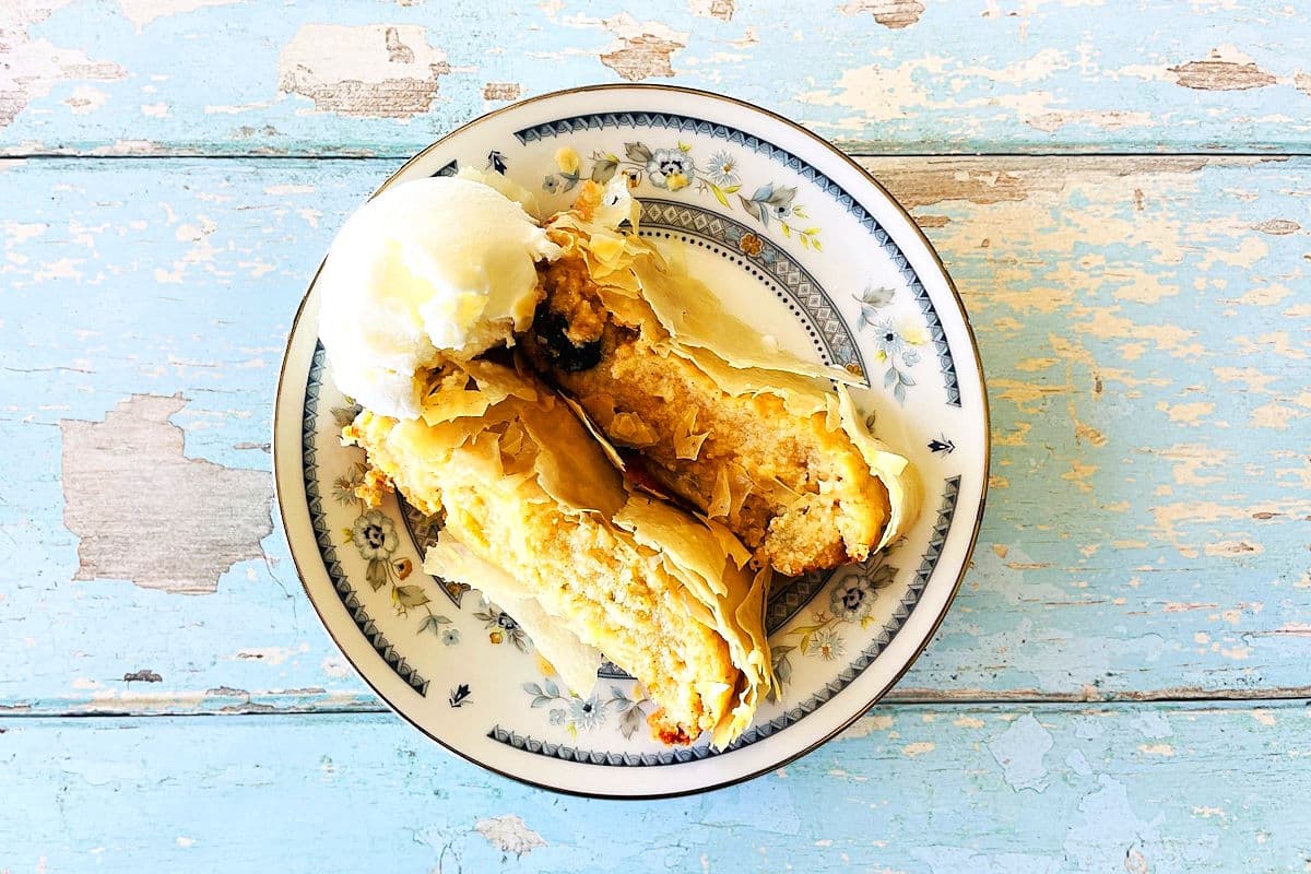 A slice of pastry dessert with a scoop of vanilla ice cream on a decorative plate, placed on a weathered blue wooden table.