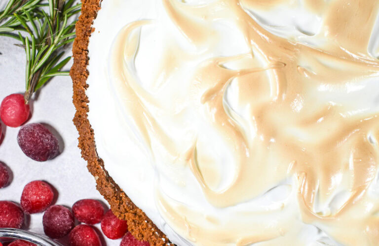 Close-up of a pie with browned meringue topping, garnished with rosemary and cranberries on the side.