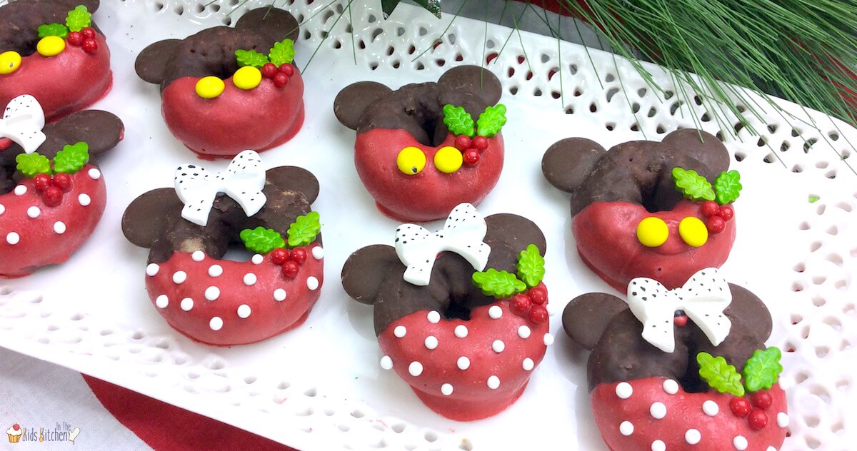 Mini donuts decorated with red and chocolate icing to resemble Mickey and Minnie Mouse, featuring candy bows, holly leaves, and yellow button details, arranged on a white platter.