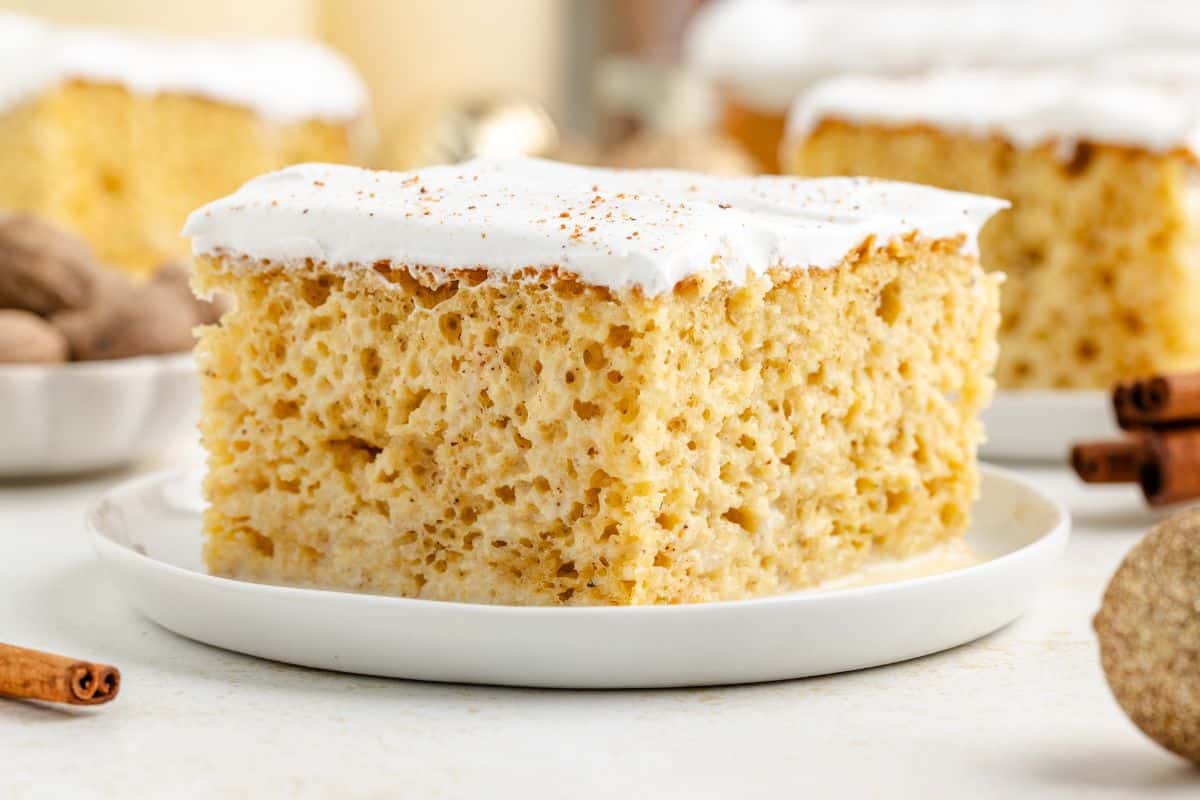 A square slice of spiced cake with white frosting on top, served on a white plate with cinnamon sticks and nutmeg nearby.