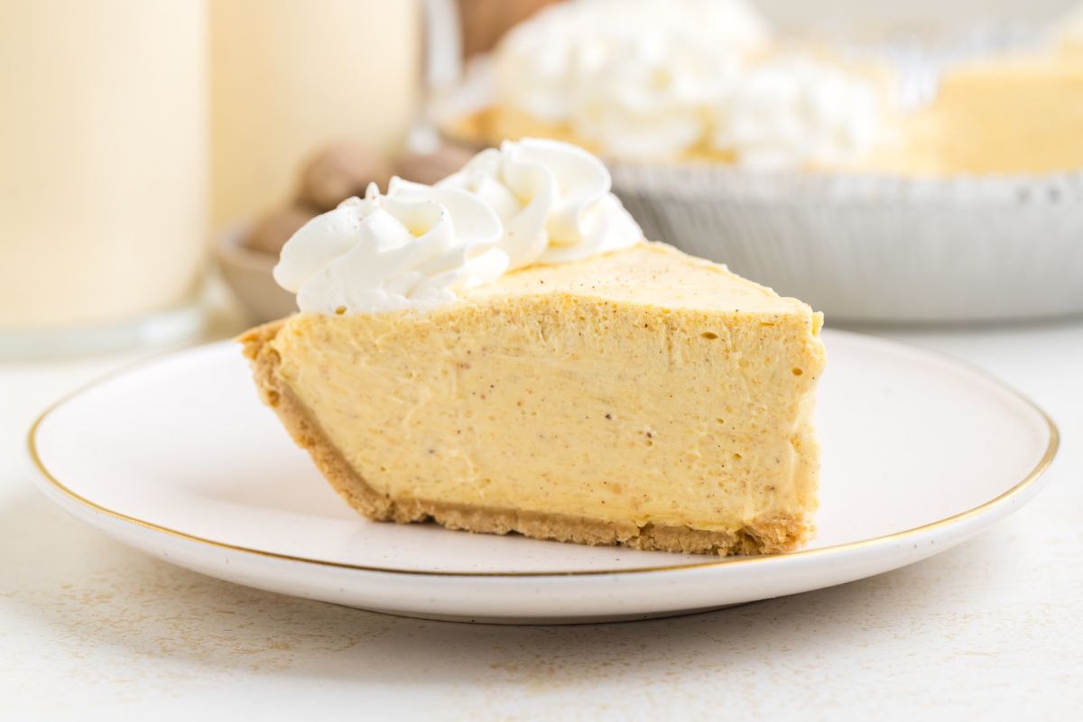 A slice of creamy pie with a graham cracker crust, topped with whipped cream, sits on a white plate.