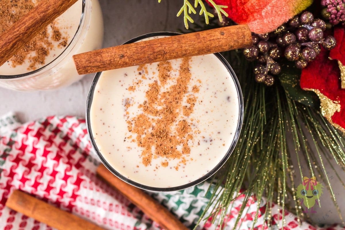A glass of eggnog topped with ground nutmeg and a cinnamon stick, surrounded by festive holiday decorations and a red-and-green cloth.
