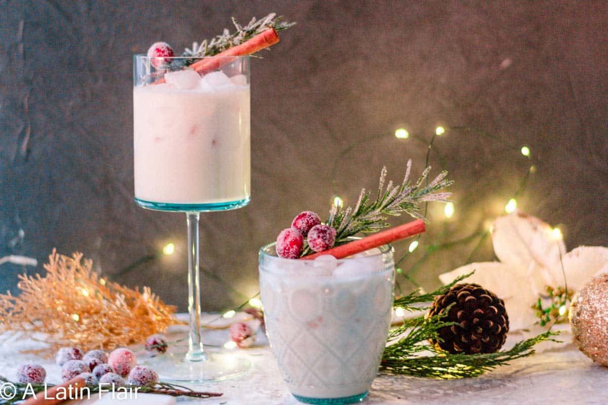 Two glasses of a creamy drink with ice, garnished with sugared cranberries, rosemary sprigs, and cinnamon sticks. Festive decorations and string lights are in the background.