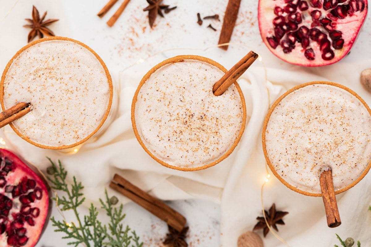 Three glasses of eggnog topped with ground nutmeg, each garnished with a cinnamon stick, surrounded by cinnamon sticks, star anise, pomegranate halves, and greenery.
