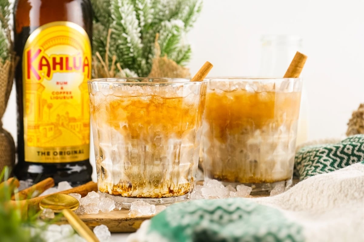 Two glasses of iced cocktails with Kahlua liqueur and cinnamon sticks, set on crushed ice with a Kahlua bottle and festive decorations in the background.