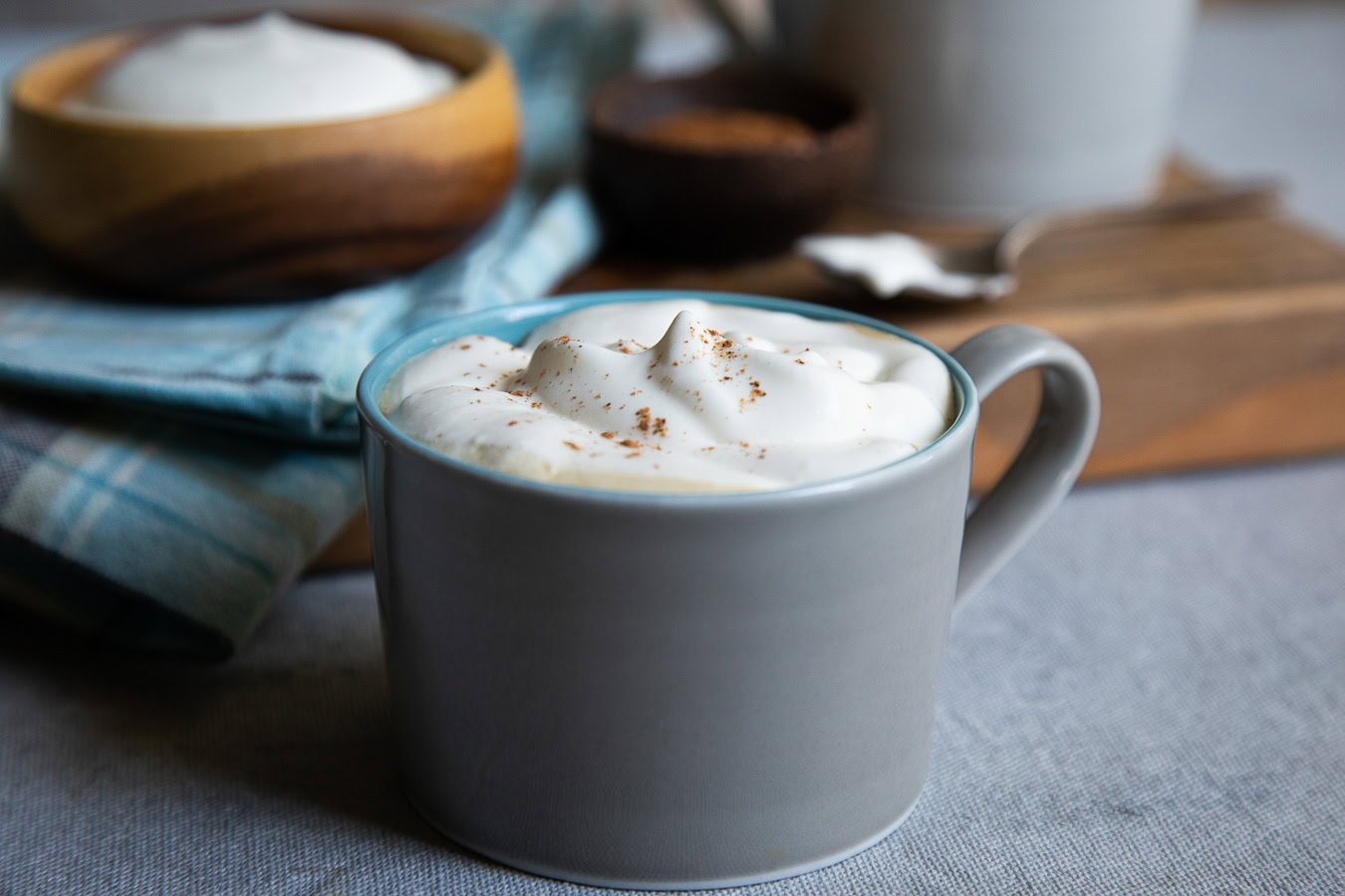 A grey mug filled with a creamy beverage topped with whipped cream sits on a table, with a blue plaid napkin and wooden bowl in the background.
