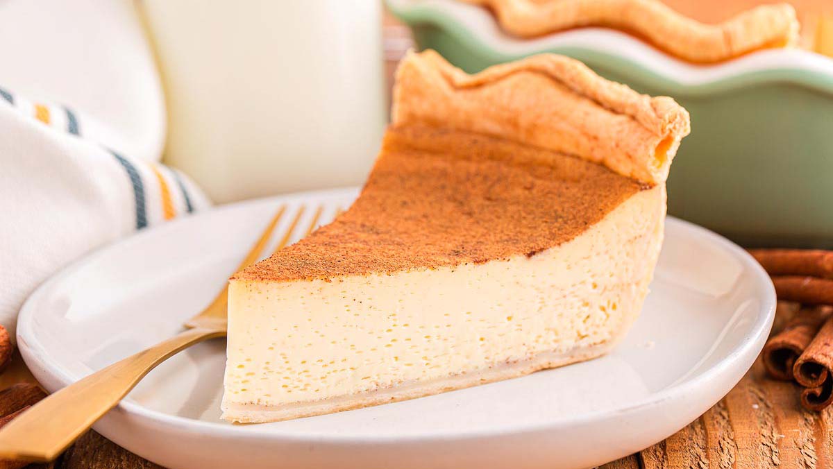A slice of custard pie with a golden crust and sprinkled cinnamon sits on a white plate next to a fork.